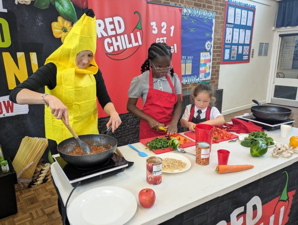 A woman in a banana costume stirs a pan on the left, while two girls in red aprons chop vegetables on a cutting board during a cooking demonstration.
