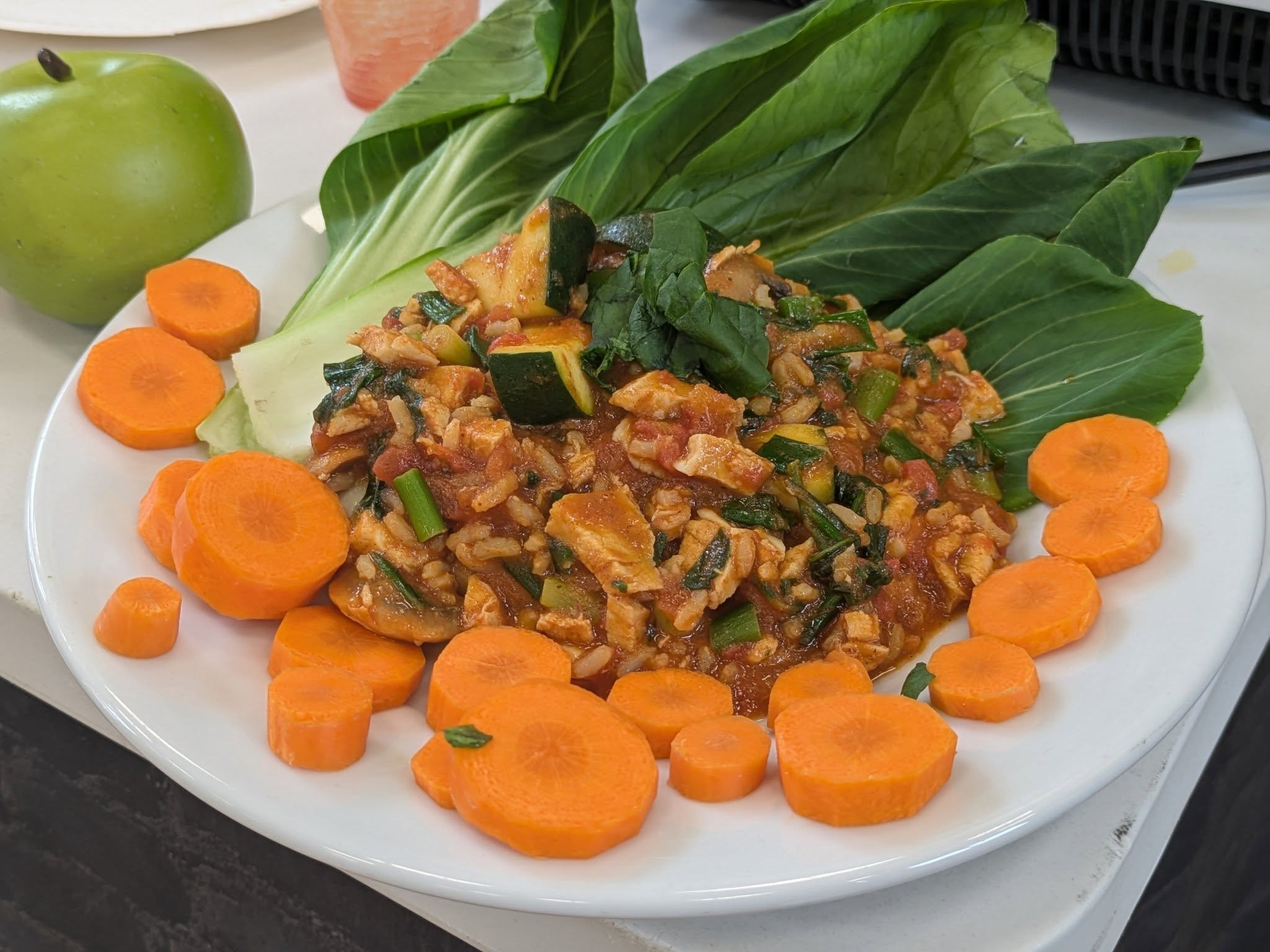 A white plate features a mound of rice and vegetable stir-fry, surrounded by sliced carrots and garnished with large bok choy or similar green leaves and a green apple on the side.