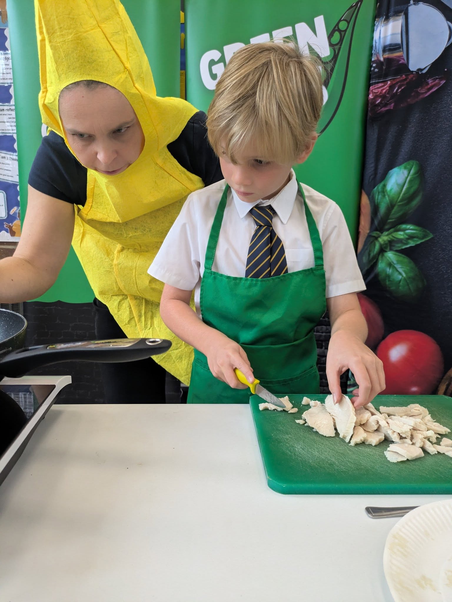 A boy in a school uniform and a green apron is chopping cooked chicken on a green cutting board, while a woman in a banana costume leans over him to supervise.