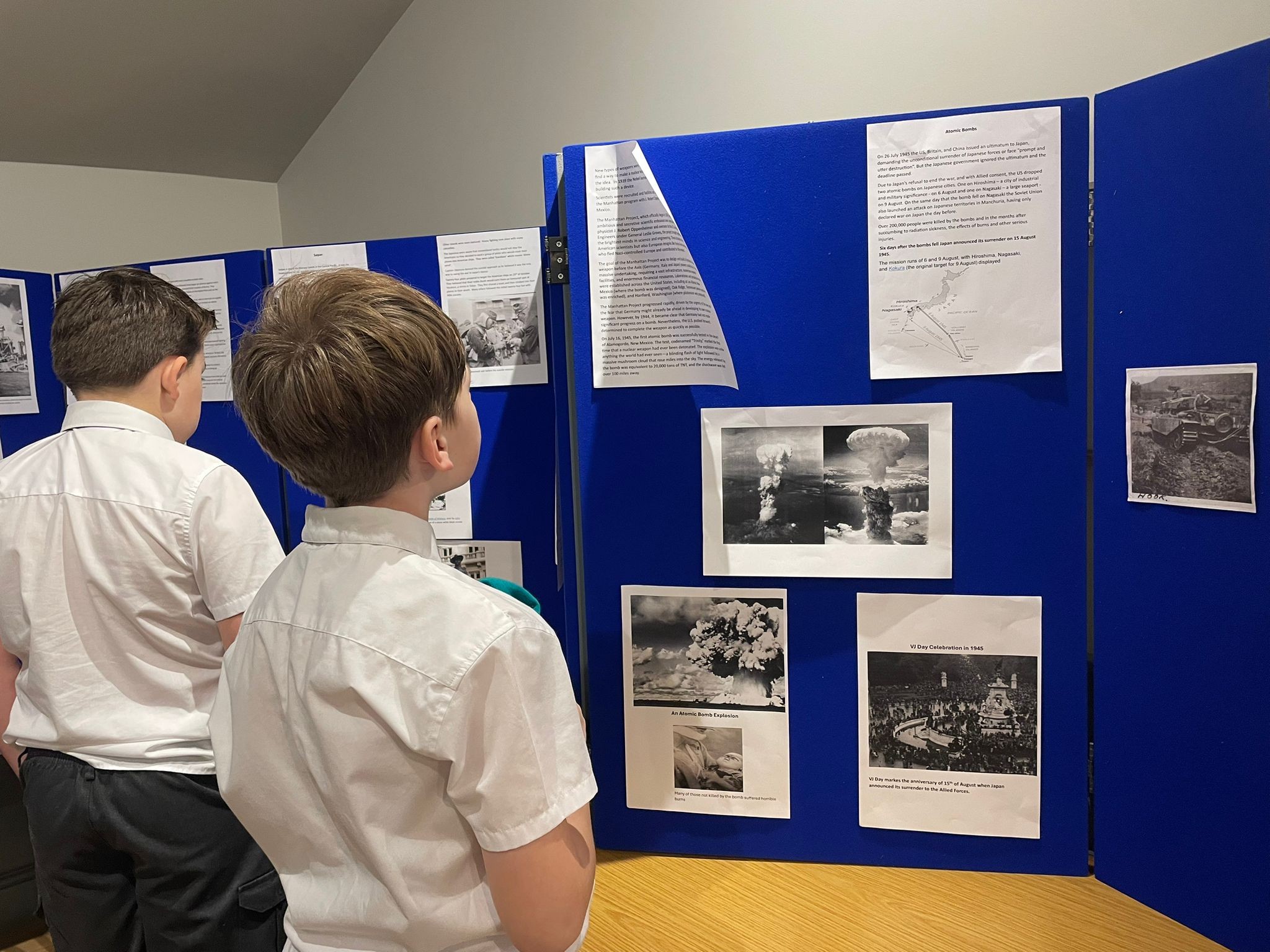 Two boys in school uniform stand looking closely at a blue display board featuring historical text and black-and-white photos related to World War II.