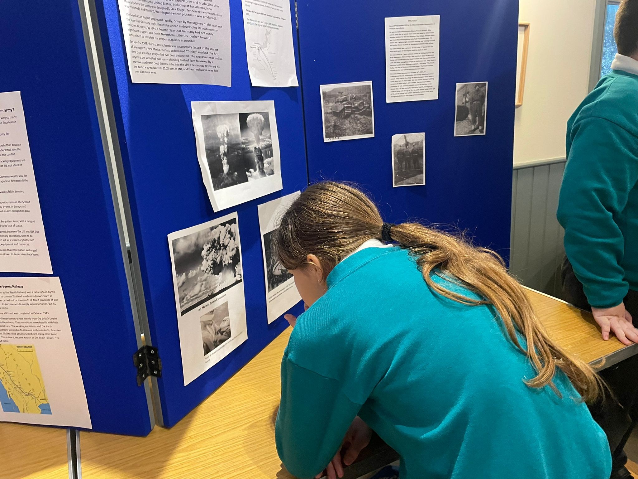 A girl with long brown hair in a teal school uniform leans close to a blue display board to examine black-and-white photos and informational text about historical events.