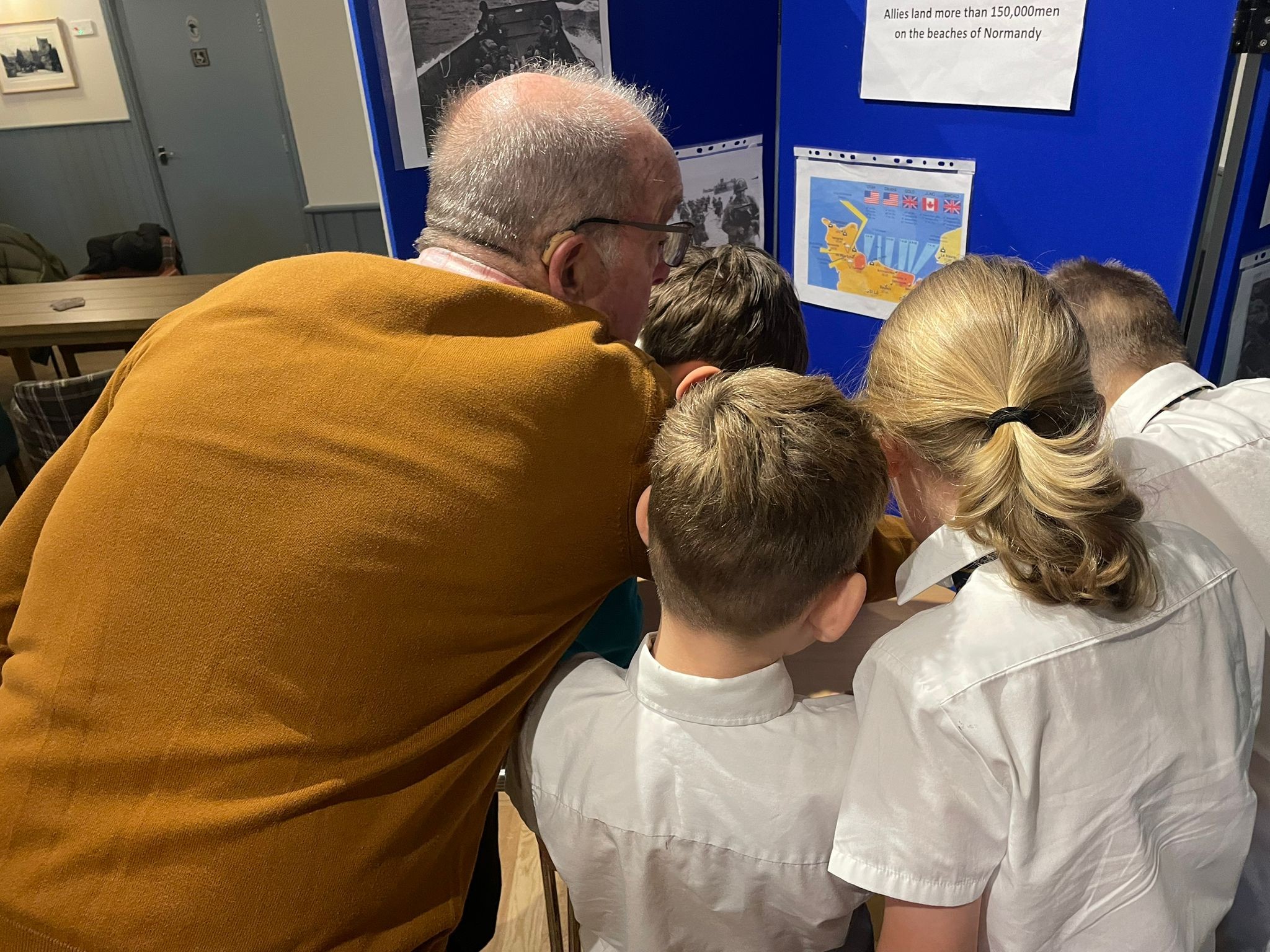 An older man wearing a brown sweater leans over to look at a blue display board with several schoolchildren, all focused on a map and photos on the board.
