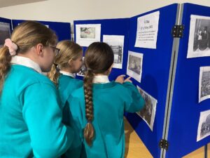 Three girls in teal school uniform stand facing a blue display board, pointing at and studying historical photos and text, including a section titled "V-E Day 8th of May 1945."