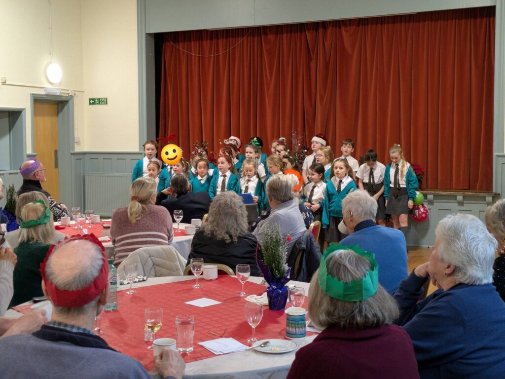 A school choir performs for a room of adults, some wearing Christmas hats